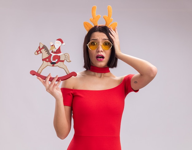 Concerned young girl wearing reindeer antlers headband and glasses holding santa on rocking horse figurine keeping hand on head looking at camera isolated on white background