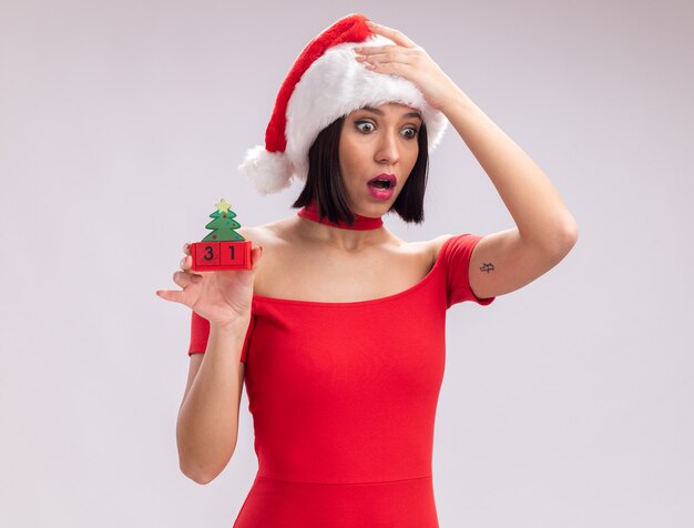 Concerned young girl wearing santa hat holding christmas tree toy with date looking down keeping hand on head isolated on white background