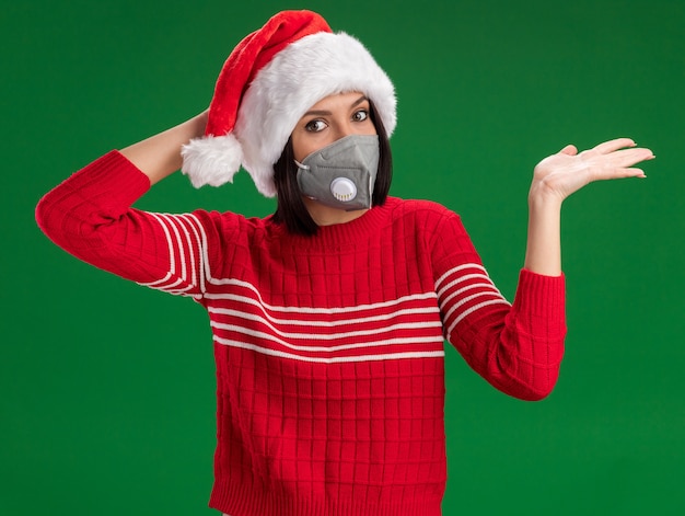 Free Photo concerned young girl wearing santa hat and protective mask looking at camera keeping hand on head showing empty hand isolated on green background