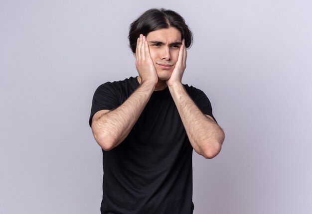 Concerned young handsome guy wearing black t-shirt putting hands on cheeks isolated on white wall