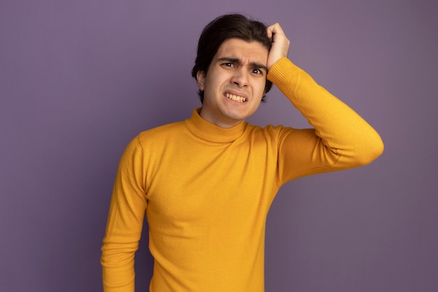 Concerned young handsome guy wearing yellow turtleneck sweater putting hand on temple isolated on purple wall