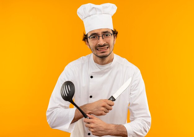 Concerned young male cook wearing chef uniform and glasses crossing hands and holding spatula with knife 