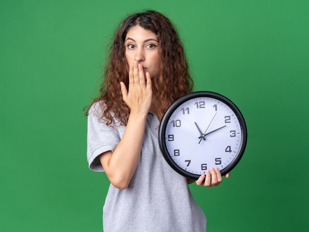 Concerned young pretty caucasian girl holding clock keeping hand on mouth  isolated on green wall with copy space