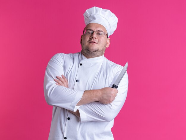 confident adult male cook wearing chef uniform and glasses standing with closed posture holding knife looking at front isolated on pink wall with copy space