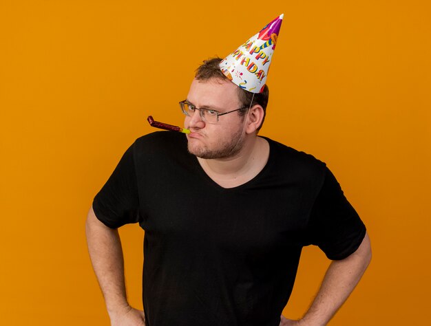 Free photo confident adult slavic man in optical glasses wearing birthday cap blowing party whistle