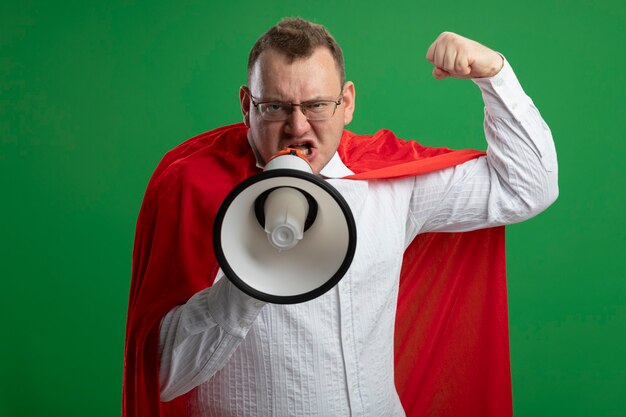 Confident adult slavic superhero man in red cape wearing glasses doing strong gesture  talking by speaker isolated on green wall