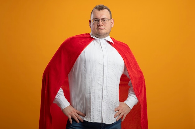 Free photo confident adult slavic superhero man in red cape wearing glasses  keeping hands on waist isolated on orange wall with copy space