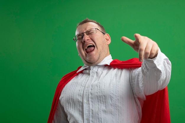 Confident adult slavic superhero man in red cape wearing glasses pointing  with closed eyes isolated on green wall with copy space