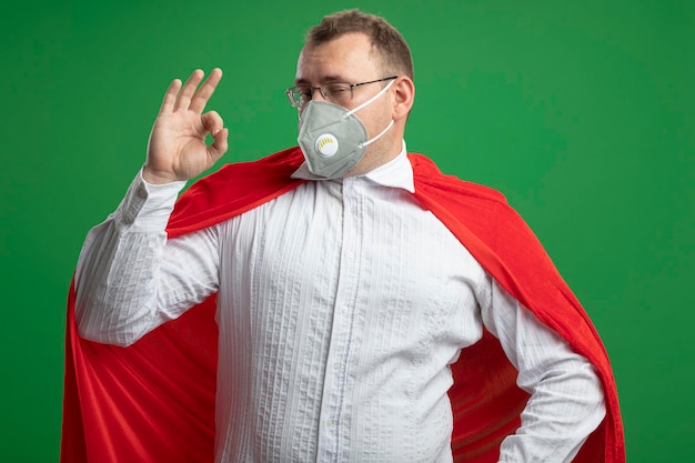 Free photo confident adult slavic superhero man in red cape wearing glasses and protective mask keeping hand on waist looking at front doing ok sign isolated on green wall