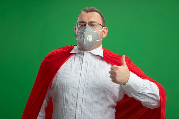 Free photo confident adult slavic superhero man in red cape wearing glasses and protective mask  showing thumb up isolated on green wall