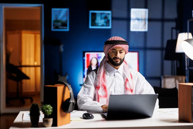 Confident Arab businessman working at a modern desk, typing on his laptop. Muslim guy engaged in browsing, email communication, and wireless connectivity in a calm and professional office setting.