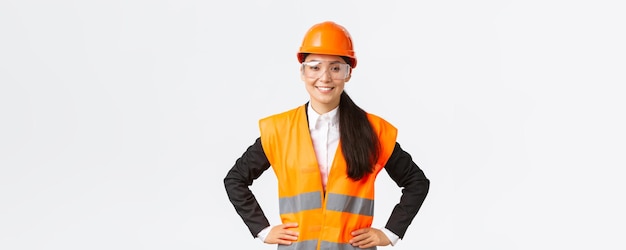 Confident asian female architect construction engineer in safety helmet protective glasses ready to enter building area for inspection smiling satisfied with results of work white background