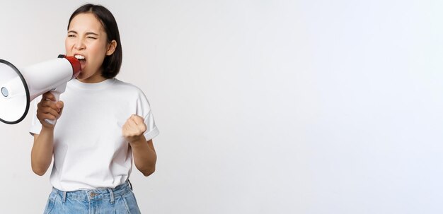 Confident asian woman shouting in megaphone screaming and protesting Girl activist using speaker to speak louder standing over white background