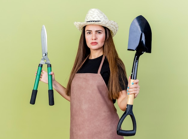 Confident beautiful gardener girl in uniform wearing gardening hat holding clippers with spade isolated on olive green background