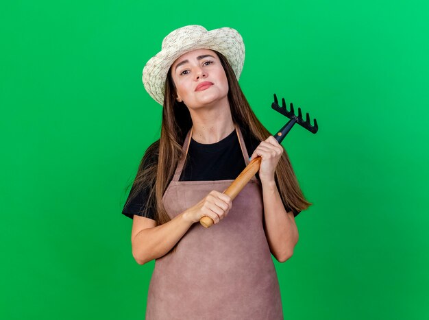 Confident beautiful gardener girl in uniform wearing gardening hat holding rake isolated on green background