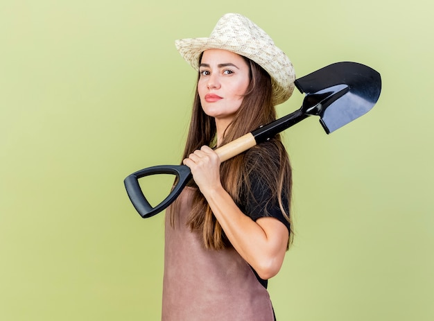 Free Photo confident beautiful gardener girl in uniform wearing gardening hat putting spade on shoulder isolated on olive green background