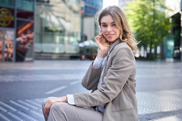 Free photo confident blond businesswoman in suit smiles at camera sits outdoors near office buildings