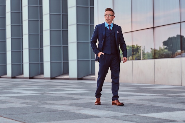 Confident businessman dressed in an elegant suit looking away and holds hand in pocket while walk on the street outdoors against cityscape background.