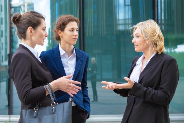 Confident businesswomen discussing project emotionally outdoors. Business colleagues wearing suits standing together in city and talking. 