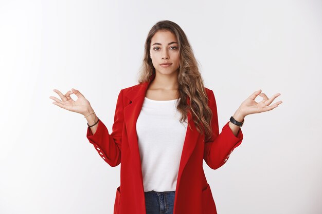 Confident calm modern successful businesswoman keeping feelings under control, showing lotus pose mudra gesture standing nirvana peacefully, stress-free, meditating standing office white wall