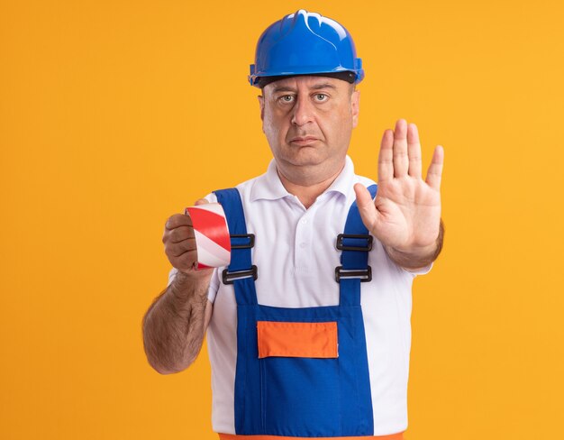 Confident caucasian adult builder man in uniform holds scotch tape and gestures stop hand sign on orange