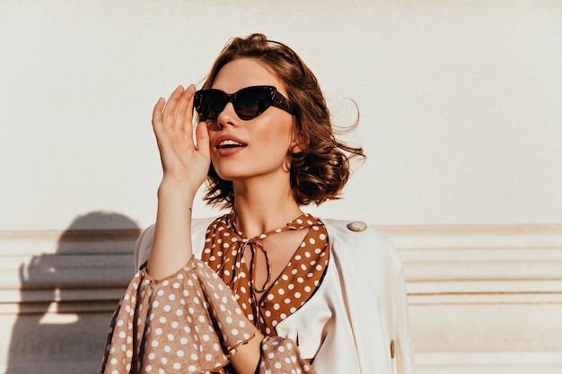 Confident caucasian girl in dark sunglasses looking in distance. Outdoor shot of good-humoured fashionable woman.