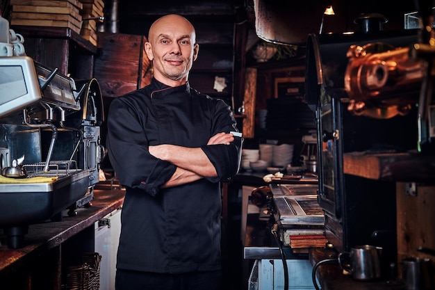 Confident chef wearing uniform posing with his arms crossed and looking at a camera in a restaurant kitchen.