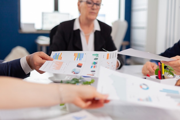 Confident company manager giving working tasks to diverse teamworkers analysing paperwork with graphs sitting in start up office