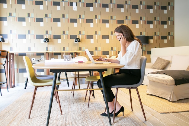 Free Photo confident creative professional discussing project with customer on phone, sitting at table with laptop and blueprints and typing