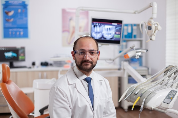 Free photo confident dentist in stomatology cabinet with orange equiptment wearing dental uniform. medical specialist in oral hygiene wearing lab coat looking at camera in dentistry office.