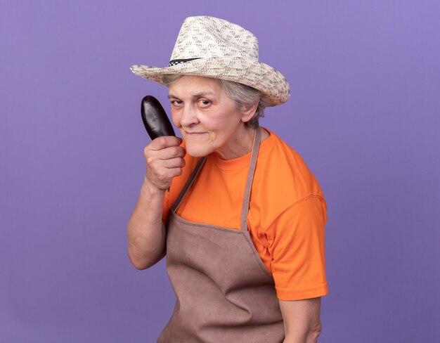 Confident elderly female gardener wearing gardening hat holding eggplant