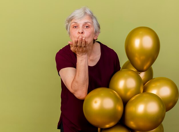 Confident elderly woman stands with helium balloons sending isolated on olive green wall with copy space