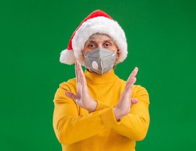 Confident elderly woman with santa hat wearing medical mask gesturing no sign with crossed hands isolated on green background with copy space