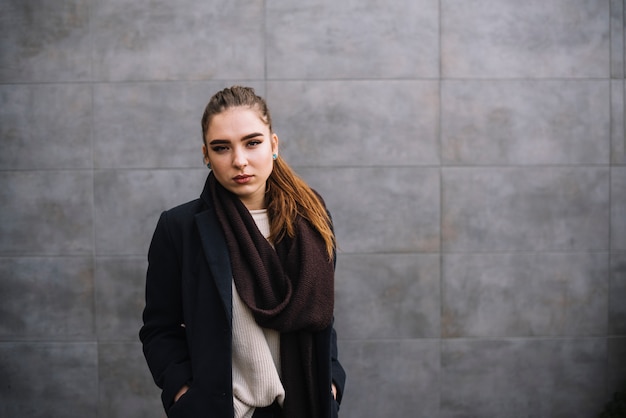 Confident elegant young woman in coat with scarf near grey wall