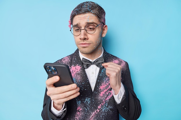 Confident European man adjustes bowtie receives messages of congratulation on anniversary dressed in festive formal suit looks attentively at smartphone screen isolated over blue background