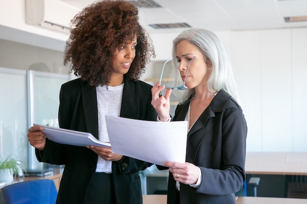 Confident female partners discussing document in office room. Two attractive successful focused businesswomen studying documentation report together. Teamwork, business and management concept