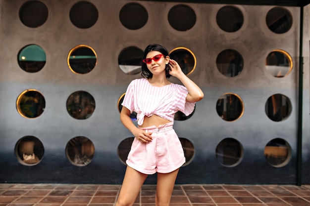 Free photo confident girl in shorts standing on urban background outdoor shot of tanned brunette woman posing near big construction