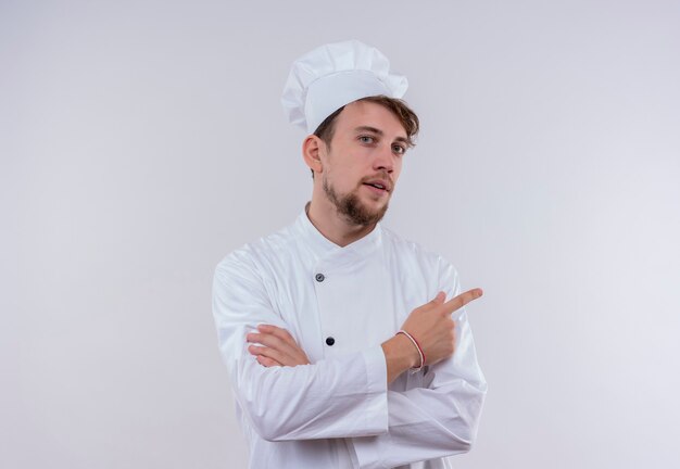 A confident handsome young bearded chef man wearing white cooker uniform and hat pointing up while looking on a white wall