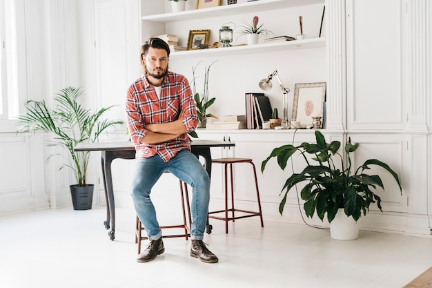 Confident handsome young man sitting on stool at home with his crossed arms