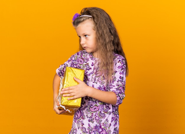 confident little blonde girl holding gift box and looking at side isolated on orange wall with copy space