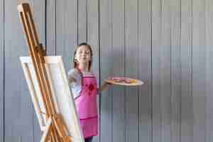 Free photo confident little girl painting on the easel standing against grey wooden wall