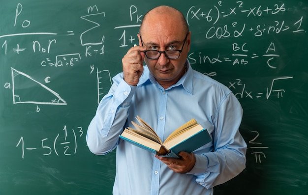 Free Photo confident looking camera middle-aged male teacher wearing glasses standing in front blackboard holding book