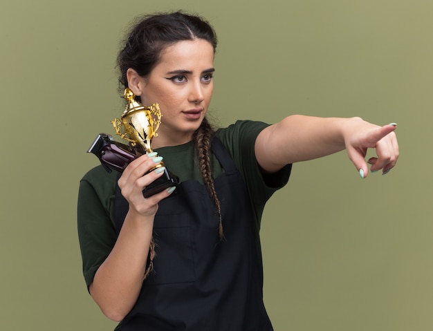 Free photo confident looking at side young female barber in uniform holding hair clippers with winner cup points at side isolated on olive green wall