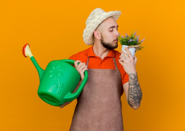 Confident male gardener wearing gardening hat holds watering can and smells flowers in flowerpot 