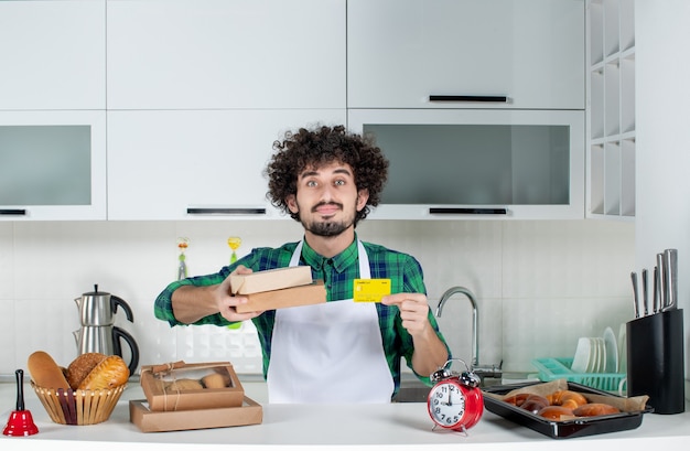 Confident man standing behind the table various pastries on it and holding bank card brown boxes in the white kitchen