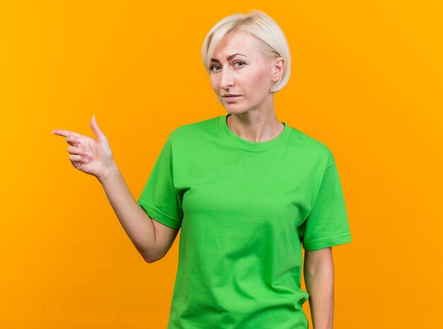 Confident middle-aged blonde slavic woman looking at camera pointing at side isolated on yellow background with copy space