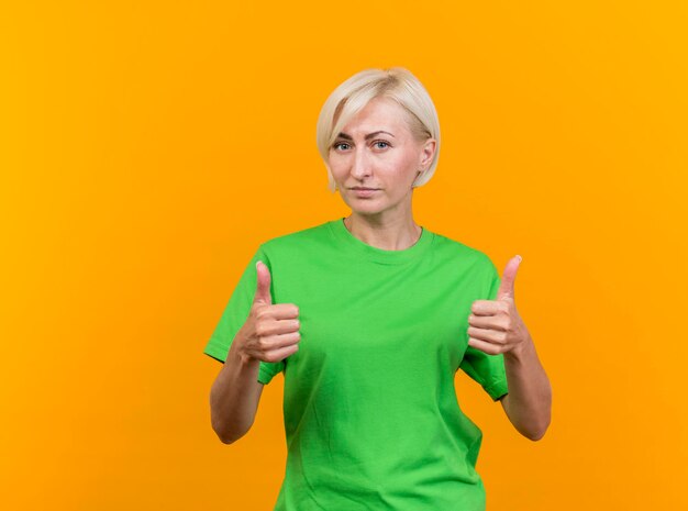 Confident middle-aged blonde slavic woman looking at front showing thumbs up isolated on yellow wall with copy space