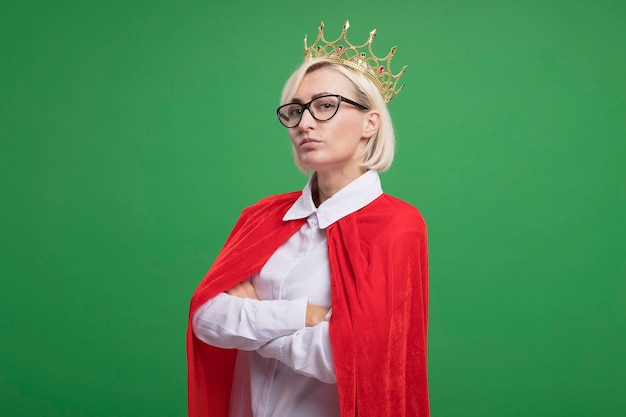 Free photo confident middle-aged blonde superhero woman in red cape wearing glasses and crown standing with closed posture in profile view