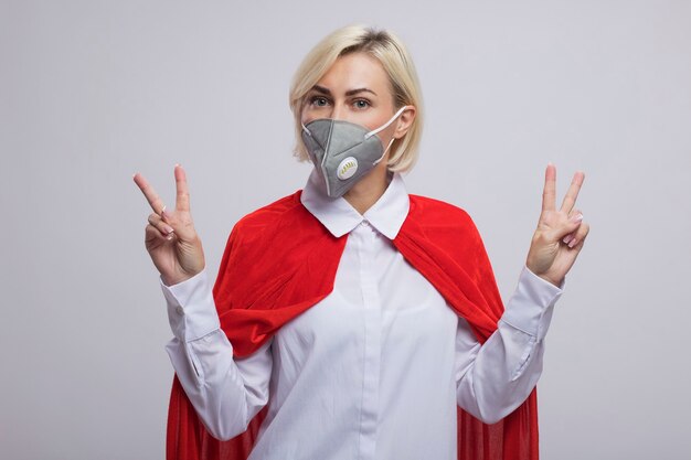 Confident middle-aged blonde superhero woman in red cape wearing protective mask  doing peace sign isolated on white wall