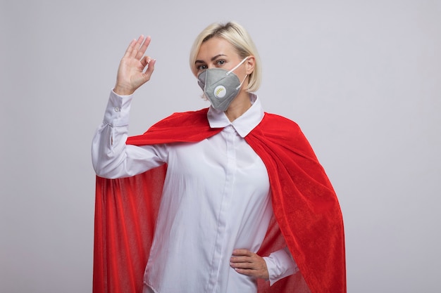 Confident middle-aged blonde superhero woman in red cape wearing protective mask keeping hand on waist  doing ok sign isolated on white wall with copy space